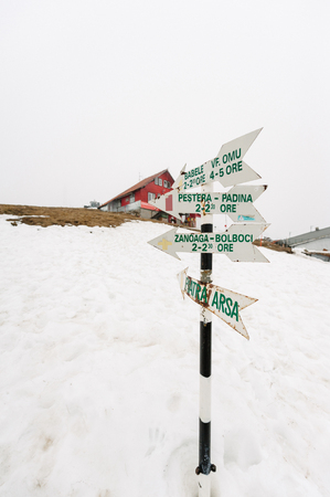 Wooden signpost in snow above Romanian ski resort village with directions and time to Varful Omu, Babele, Pestera Padina, Zanoaga Bolboci, Piatra Arsaの写真素材