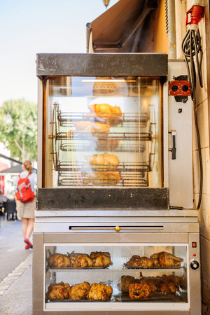 Grilled roasted chickens on a spit in rotisserie with beautiful aroma and smoke on the streets of a city with a women with backpack wlaking nearbyの写真素材