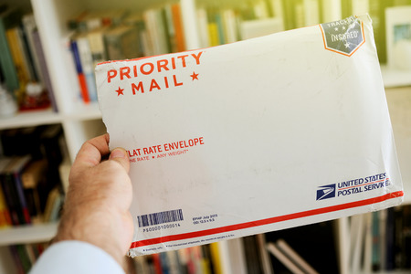 LONDON, UNITED KINGDOM - OCTOBER 18, 2014: Damaged USPS United States Postal Service Parcel envelope in man's hand displaying damaged documents on boockself background. The United States Postal Service, also known as the Post Office, U.S. Mail, or Postal のeditorial素材