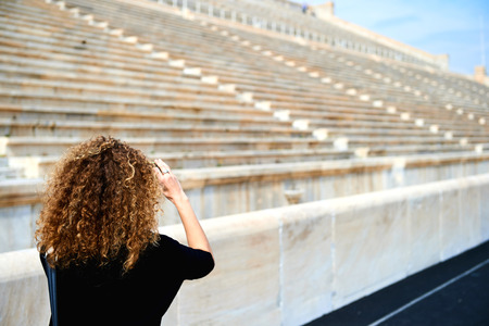 Rear view of elegant woman taking photo on smartphone outside the stadium of the ancient stone stairs chairsの写真素材