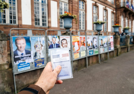 STRASBOURG, FRANCE - APR 23, 2017: Man holding Carte Electorale - voter's card French voter registration card in front of official campaign posters for all eleven candidates for the 2017 French presidential elections outside pooling stationのeditorial素材