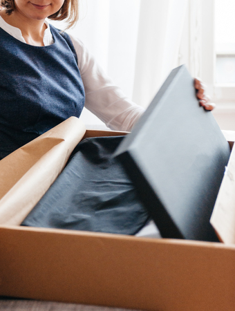 Young beautiful woman on living room sofa looks happy as she unpacks unbox cardboard box parcel containing new beautiful fashion clothes and shoesの写真素材
