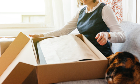 Curious cat helping young beautiful woman on living room sofa looks happy as she unpacks unbox cardboard box parcel containing new beautiful fashion clothesの写真素材
