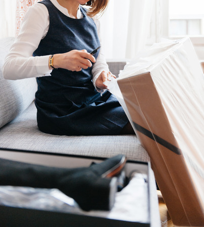 Young beautiful woman on living room sofa using cutter to unpacks unbox cardboard box parcel containing new beautiful fashion clothesの写真素材