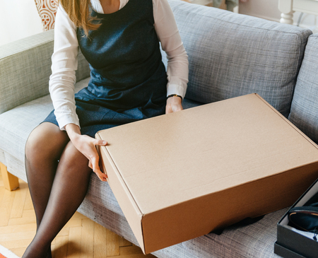 Unboxing cardboard by young beautiful woman on living room sofa - box parcel containing new beautiful fashion clothesの写真素材