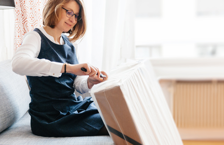 French young beautiful woman on living room sofa looks happy as she unpacks unbox cardboard box parcel containing new beautiful fashion clothesの写真素材