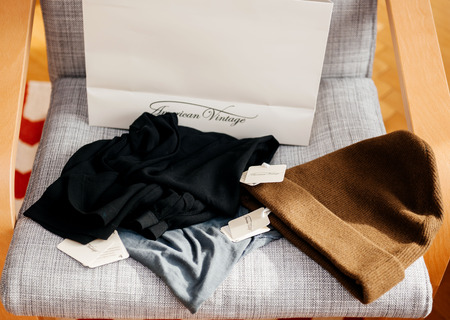 PARIS, FRANCE - OCT 24, 2016: White shopping bag with AMERICAN VINTAGE fashion brand logotype on the armchair with multiple accesories presentatin, wool hat, t-shirt, shirtのeditorial素材