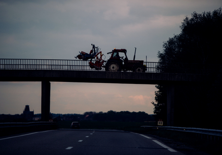 Silhouete of agricultural tractor with special trailer for harvesting driving on the bridge above fast German autobahnの写真素材