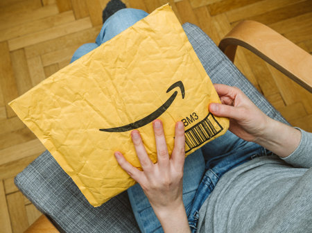 PARIS, FRANCE - NOV 4, 2017: Living room view from above of woman in armchair unpacking unboxing Amazon cardboard envelope with smile logotype printedのeditorial素材