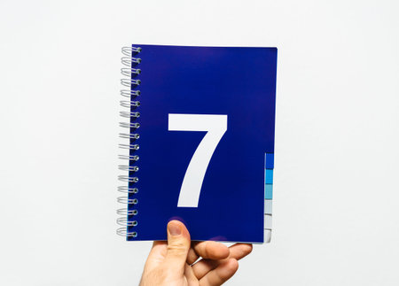 Male hand holding notebook with big number 7 printed on blue color ready for presentation or...