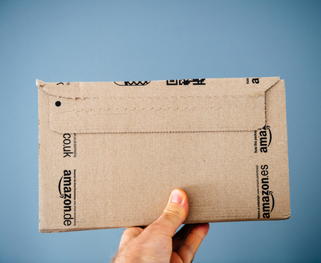 PARIS, FRANCE - APR 24, 2018: Man holding rear part of the Amazon Prime cardboard delivery box against gray background - on-line e-commerce shoppingのeditorial素材