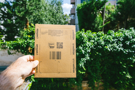 PARIS, FRANCE - NOV 28, 2017: Male hand holding the Amazon cardboard box as he picked it from the front door with trees and houses in the backgroundのeditorial素材