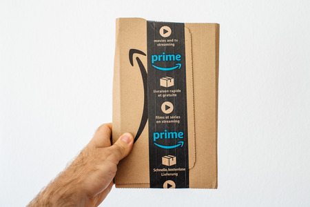PARIS, FRANCE - APR 16, 2018: Man holding on vertical position against white background Amazon Prime cardboard box with diverse  in multiple languages on scotch band - showing the fast delivery boxのeditorial素材