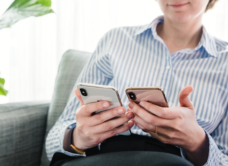 PARIS, FRANCE - SEP 27, 2018: Beautiful French woman testing and comparing the newest latest iPhone Xs and Xs Max smartphone telephone from Apple Computers on office living room sofaのeditorial素材