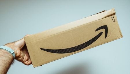 Paris, France - Jun 11, 2018: Man hand holding new freshly delivered Amazon Prime cardboard box featuring big smile blue backgroundのeditorial素材