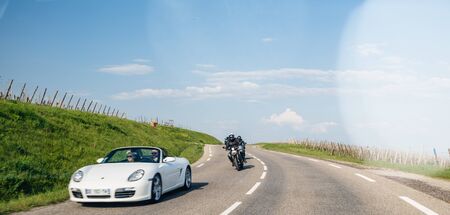 Bergheim, France - Apr 19, 2019: Group of motorcyclists following couple driving in white luxury convertible Porsche sport car driving fast on Alsatian village roadのeditorial素材