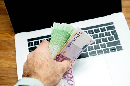 Man hand holding large stack pile of 500 100 200 Euro bank currency notes above the keyboard of new laptop computer keyboard on wooden tableの写真素材