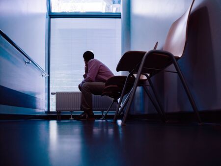 Low angle view of lonely patient sitting in modern hospital waiting lobby room looking through window with blinds as he waits for good or bad news from his doctorの写真素材