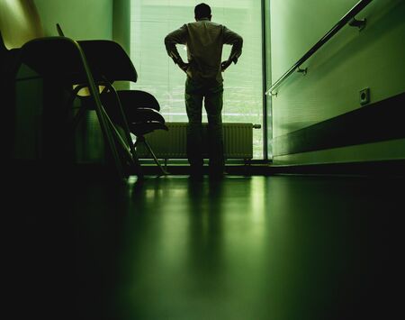 Low angle view of lonely patient in full length in modern hospital waiting lobby room uin front of window hands on hips as he waits for good or bad news from his doctorの写真素材
