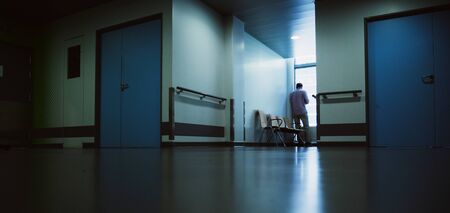 Lonely patient in full length in modern hospital waiting lobby room near the window as he waits for good or bad news from his doctor - blue hospital atmosphereの写真素材