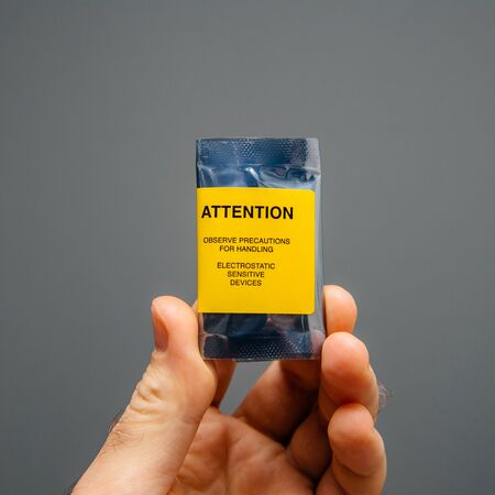 Man hand holding against gray background tiny plastic package with PC component and yellow sticker Attention Static Sensitive Devicesの写真素材