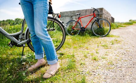 Feets of adult woman in blue jeans denim making a pause after riding the bike on gravel road with orange bike in the backgroundの写真素材