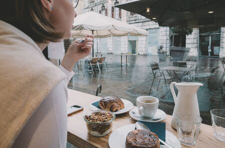 Rear view of single woman eating delicious yogurt with muesli drinking coffee with croissant and cake early in the morning in French cafe with overlook to central square terraceの写真素材