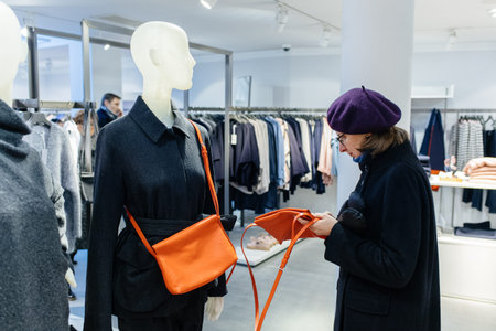 Beautiful French woman wearing purple beret hat looking at the thin orange leather purse in fashionable clothing storeの写真素材