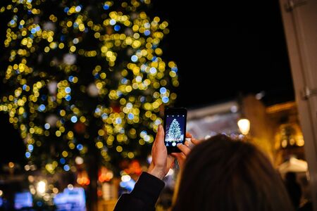 Strasbourg, France - Nov 23, 2017: Rear view of adult woman rising smartphone to take a photograph on smartphone the illuminated and decorated Christmas fir tree at the market capitalのeditorial素材