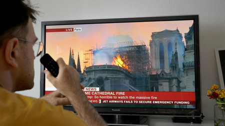 Paris, France - April 15, 2019: Man holding remote control looking at the TV in living room breaking last news with British BBC World News channel live broadcast from the Notre-Dame de Paris fireのeditorial素材