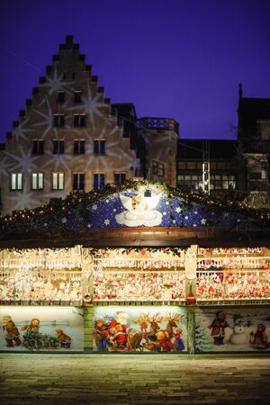 Strasbourg, France - Dec 20, 2016: Market stall chalet with multiple toys for sale and seller in background arranging toys and illuminated Christmas decorated Alsatian build in backgroundのeditorial素材