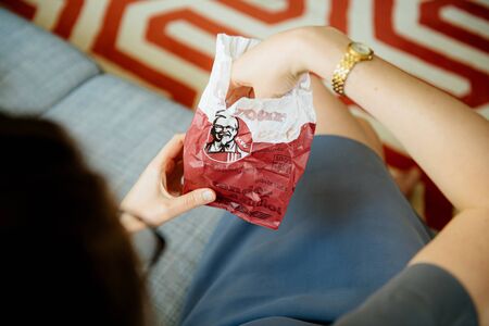 Paris, France - Aug 1, 2019: Woman taking from the KFC meat paper bag fresh delicious meat - overhead view in living room couchのeditorial素材