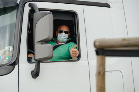 Kehl, Germany - Mar 16, 2020: Truck driver in Mercedes-Benz Actros wearing a protection mask shows...