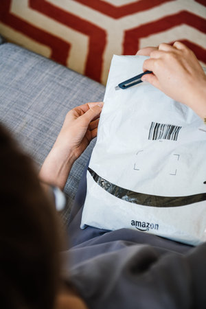 Paris, France - Aug 1, 2019: Overhead view of elegant woman on living room couch holding open with cutter fresh Amazon Prime plastic package parcel opening with cutterのeditorial素材