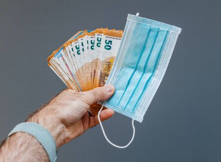 Man hand holding surgical procedure medical face mask with thousands of money banknotes euros during worldwide shortage of masks - as demand during quarantine in US, Europe, Asia during global pandemicの写真素材