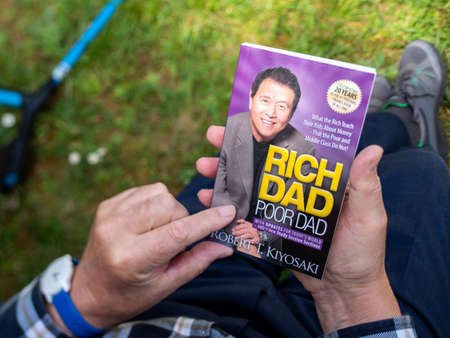 Paris, France - May 3, 2020: Overhead POV view of senior male hand hodling in had reading the iconic book Rich dad and Poor Dad by Robert T Kiyosakiのeditorial素材