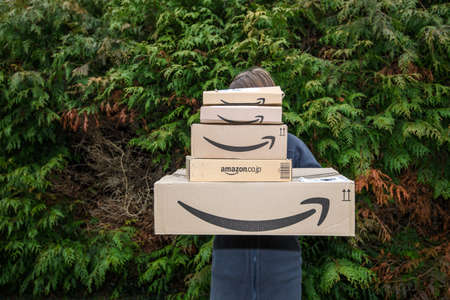 Paris, France - Feb 23, 2020: Front view of young delivery woman holding multiple a huge stack of Amazon prime cardboard parcels cardboard boxes with one of it coming from Japanのeditorial素材