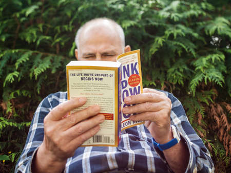 Paris, France - May 3, 2020: Front view of senior curious male reading Think and grow rich book by Napoleon Hill in green gardenのeditorial素材