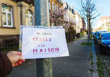 Strasbourg, France - April 2020: POV male hand holding paper De Grace restez a la maison - translated as Stay at home message painted by kid sticked on pillar during coronavirus covid-019 pandemicのeditorial素材