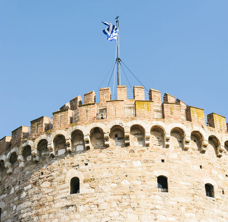 White Tower of Thessaloniki with waving flag against clear blue