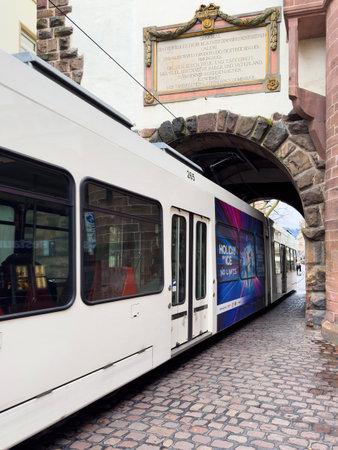 Freiburg, Germany - Mar 5, 2024: A modern tram passes through a historic stone archway in Freiburg im Breisgau. The arch is adorned with a commemorative plaque, blending contemporary transit with historical architectureのeditorial素材