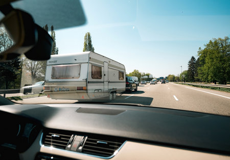 Alsace, France - Apr 19, 2019: A car towing a caravan on a busy highway, captured from inside another vehicle, with trees and a clear sky in the background.のeditorial素材
