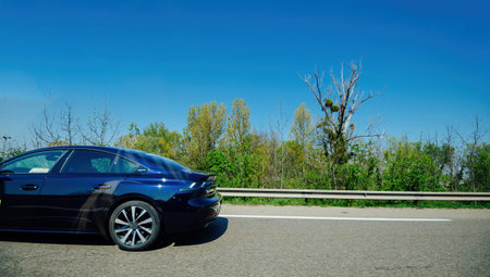 A blue sedan car driving along a highway with a clear blue sky and greenery in the background,...