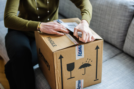 Paris, France - Oct 20, 2015: Woman opening a fragile Amway package on a grey couch, carefully unsealing the box to reveal its contents, highlighting the process of receiving a deliveryのeditorial素材