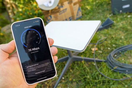 Germany - Jun 15, 2023: Hand holding a smartphone displaying a speed test result of 70 Mbps with a satellite dish and outdoor setup visible in the background amidst green surroundingsのeditorial素材