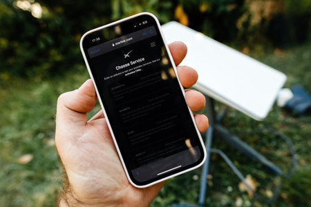 Germany - Jun 15, 2023: A hand holding a smartphone displaying the Starlink service selection page with a satellite dish in the background, amidst a green outdoor setting.のeditorial素材