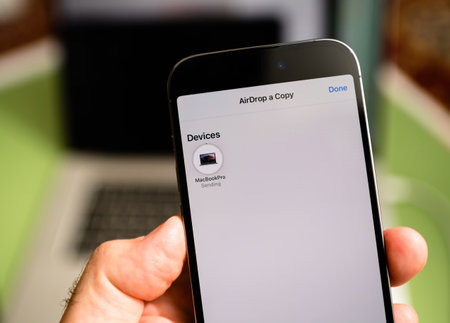 Paris, France - Sep 29, 2022: A hand holding an iPhone by Apple Computers displaying the AirDrop interface with a MacBook Pro in the background, illustrating the process of wirelessly sending a file between Apple devices in a modern tech settingのeditorial素材