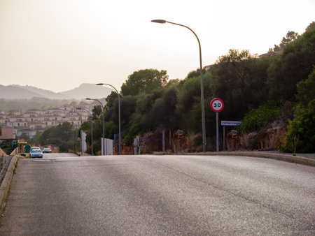 A scenic view of a road in SEstany den Mas, Mallorca, lined with street lamps and a 30 km h speed limit sign, with residential houses and hills in the background during sunsetの写真素材