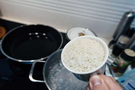 POV of a male hand adding a cup of basmati rice to boiling water in a kitchen, highlighting the cooking process as steam rises from the pot, emphasizing a hands-on meal preparationの写真素材