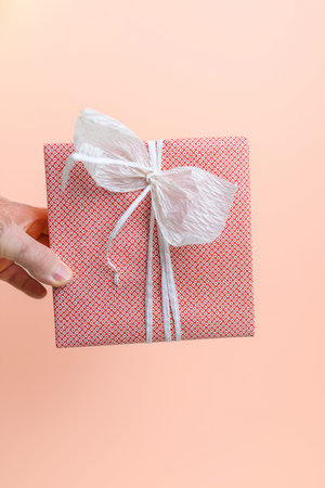 A hand holds a beautifully wrapped red gift with white ribbon against a soft peach background,...